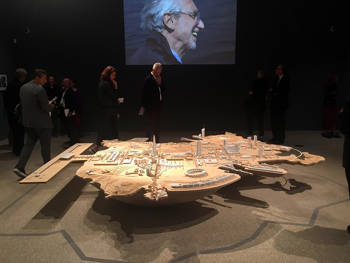 A large irregular model on which there are different kinds of buildings in the centre of an exhibition and people around are observing it
